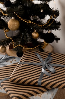 A decorated Christmas tree adorned with black and gold baubles and golden garlands. Below the tree are neatly wrapped gifts featuring black and gold striped wrapping paper and silver ribbons. The scene captures a festive and elegant holiday setting.
