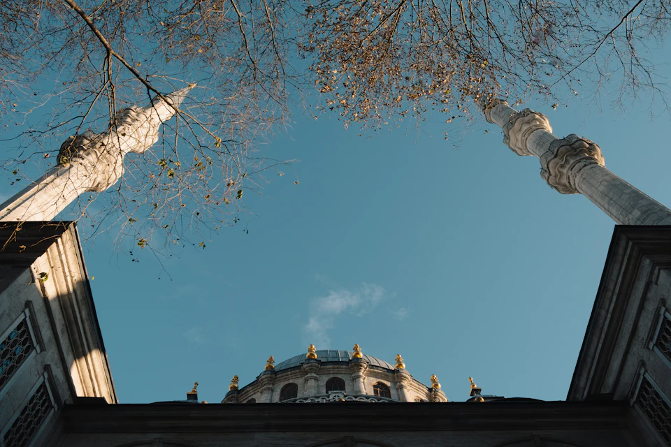 The image depicts a majestic mosque with two towering minarets, one on each side, reaching towards the clear blue sky. The mosque's dome is adorned with gold accents and stands out against the backdrop of the azure sky. The perspective of the photo is from below, looking up at the minarets, emphasizing their height and grandeur.

The image also features a few trees scattered around the scene, adding a touch of nature to the architectural marvel.