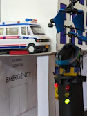 A model scene depicting an emergency hospital setting with an ambulance vehicle and a helicopter landing on a rooftop. The models are detailed with an 'Emergency' sign and lights on the helicopter. The backdrop includes a hospital facade.