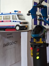 A model scene depicting an emergency hospital setting with an ambulance vehicle and a helicopter landing on a rooftop. The models are detailed with an 'Emergency' sign and lights on the helicopter. The backdrop includes a hospital facade.