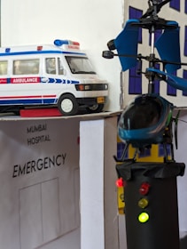 A model scene depicting an emergency hospital setting with an ambulance vehicle and a helicopter landing on a rooftop. The models are detailed with an 'Emergency' sign and lights on the helicopter. The backdrop includes a hospital facade.