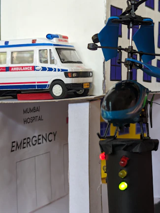 A model scene depicting an emergency hospital setting with an ambulance vehicle and a helicopter landing on a rooftop. The models are detailed with an 'Emergency' sign and lights on the helicopter. The backdrop includes a hospital facade.