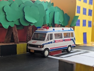 A miniature model of an ambulance is positioned on a road made from craft materials, featuring a black and yellow curb. In the background, there are stylized trees with green paper leaves and a yellow building with blue windows and door, all resembling a handcrafted diorama.