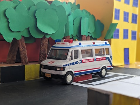 A miniature model of an ambulance is positioned on a road made from craft materials, featuring a black and yellow curb. In the background, there are stylized trees with green paper leaves and a yellow building with blue windows and door, all resembling a handcrafted diorama.