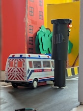 A model scene features a toy ambulance on a roadway next to a tall, cylindrical black traffic light. The backdrop includes paper cutouts of trees and a red building with windows, contrasted against a bright yellow wall.