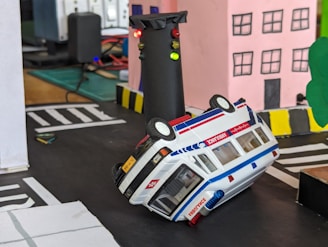 A toy ambulance is shown upside down on a makeshift road near a model traffic signal with red, yellow, and green lights. The scene includes hand-crafted model buildings and streets, suggesting a miniature diorama or road safety demonstration.