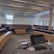 A modern lecture hall or classroom featuring tiered seating with wooden desks and blue cushioned seats. At the front, there is a lectern equipped with a monitor, a control panel, and various stationery items. The room is well-lit by natural light coming through large windows along the right side of the room. Ceiling-mounted projectors and speakers are also visible.