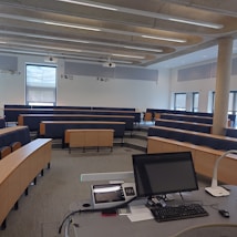 A modern lecture hall or classroom featuring tiered seating with wooden desks and blue cushioned seats. At the front, there is a lectern equipped with a monitor, a control panel, and various stationery items. The room is well-lit by natural light coming through large windows along the right side of the room. Ceiling-mounted projectors and speakers are also visible.