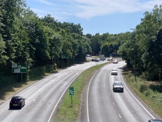 A roadway scene showing a two-lane road, bordered by lush green trees on both sides. Several vehicles, including cars and a truck, are seen traveling in both directions. Traffic lights are visible at a junction further down the road. Clear blue sky is visible above the trees.
