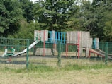 A playground with a variety of equipment made from metal and wood sits within a fenced area. The playground features structures such as slides, climbing frames, and a spring rider, all surrounded by lush, green trees and grass.
