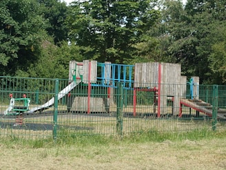 A playground with a variety of equipment made from metal and wood sits within a fenced area. The playground features structures such as slides, climbing frames, and a spring rider, all surrounded by lush, green trees and grass.