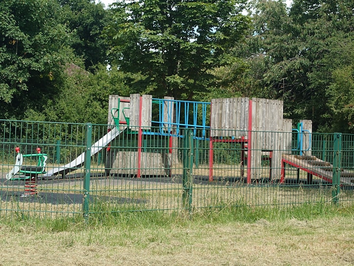 A playground with a variety of equipment made from metal and wood sits within a fenced area. The playground features structures such as slides, climbing frames, and a spring rider, all surrounded by lush, green trees and grass.
