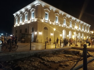 A large, illuminated classical building with arched windows and decorative columns dominates the scene. The area in front is bustling with people walking, socializing, and observing the surroundings. Streetlights cast a warm glow over the cobblestone pavement. In the foreground, there's an archaeological excavation site with exposed ancient stones.