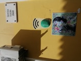 A yellow wall features a white street sign with 'Travessa Teixeira Junior' written on it. Below, there's a green brain sculpture adjacent to black curved lines resembling a Wi-Fi signal. Next to it, a colorful piece of art depicts a woman whose face is overlaid with floral and leaf patterns, framed by blue and green decorative tiles. At the bottom left, part of an air conditioning unit is visible.