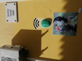 A yellow wall features a white street sign with 'Travessa Teixeira Junior' written on it. Below, there's a green brain sculpture adjacent to black curved lines resembling a Wi-Fi signal. Next to it, a colorful piece of art depicts a woman whose face is overlaid with floral and leaf patterns, framed by blue and green decorative tiles. At the bottom left, part of an air conditioning unit is visible.