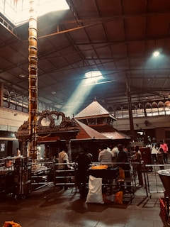 A dimly lit interior of a temple with a focus on a tall, ornate pillar under a high ceiling. Sunlight streams in through a window above, illuminating the pillar and casting light across the temple floor. People are gathered around the area, engaged in various activities, with some observing the central structure made of intricately carved wood and metal.