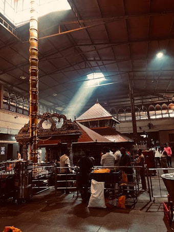 A dimly lit interior of a temple with a focus on a tall, ornate pillar under a high ceiling. Sunlight streams in through a window above, illuminating the pillar and casting light across the temple floor. People are gathered around the area, engaged in various activities, with some observing the central structure made of intricately carved wood and metal.