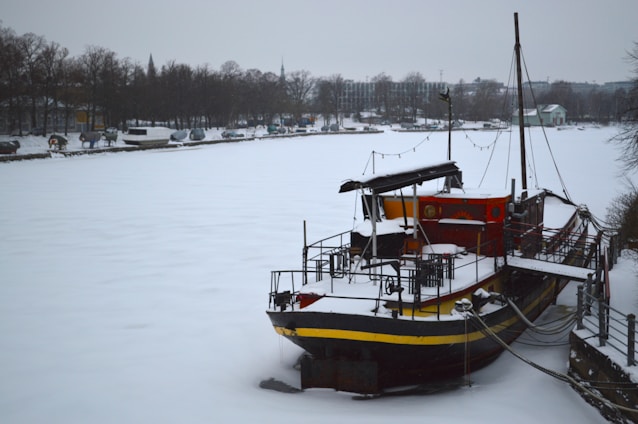 A boat is moored on a snow-covered river, with its deck and surrounding area blanketed in white snow. The background features a row of leafless trees and buildings, under a grey, overcast sky.