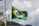 A Brazilian flag waves on a flagpole, showcasing its iconic green, yellow, and blue colors with a circular emblem in the center. The background features a cloudy sky, with some trees and a telecommunications tower in the distance.