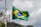 A Brazilian flag waves on a flagpole, showcasing its iconic green, yellow, and blue colors with a circular emblem in the center. The background features a cloudy sky, with some trees and a telecommunications tower in the distance.