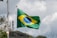 A Brazilian flag waves on a flagpole, showcasing its iconic green, yellow, and blue colors with a circular emblem in the center. The background features a cloudy sky, with some trees and a telecommunications tower in the distance.