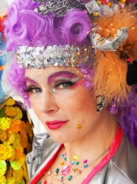 A person with colorful, elaborate makeup and costume, featuring bright purple hair, sequins, and feathers. Vibrant makeup highlights the eyes and lips, and a variety of colorful gems adorn the outfit.