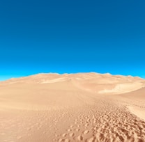 A vast desert landscape with rolling sand dunes under a clear, bright blue sky. Footprints are visible on the sandy surface, suggesting recent human activity. The image conveys a sense of openness and tranquility.