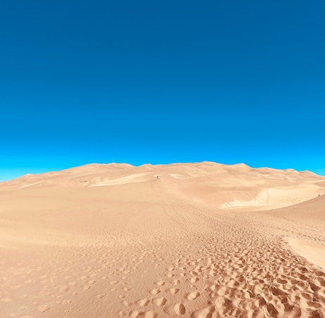 A vast desert landscape with rolling sand dunes under a clear, bright blue sky. Footprints are visible on the sandy surface, suggesting recent human activity. The image conveys a sense of openness and tranquility.
