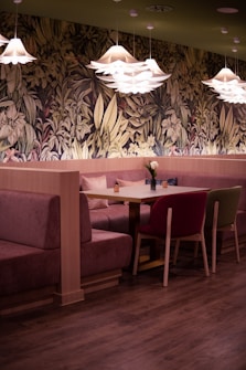 A cozy dining area features a wooden table surrounded by cushioned booths and chairs in a warm, inviting environment. The background is adorned with a detailed, nature-inspired wallpaper showcasing various leaves and flowers. The ceiling has several modern, flower-shaped pendant lights casting a soft glow, adding to the tranquil ambiance. The flooring is a dark, polished wood that complements the earthy tones.