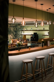 A modern bar setting featuring a row of high stools along a wooden counter. Several illuminated light bulbs hang from the ceiling, and behind the bar, there is a selection of bottles on display. The walls have a green hue with decorative leafy patterns, adding a touch of nature-inspired design.