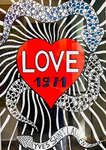 A vibrant illustration features a large red heart with the word 'LOVE' and the year '1971' prominently displayed in bold white lettering. The background includes a black and white striped pattern intertwined with a snake-like design made up of blue and white scales. At the bottom, a ribbon with the text 'YVES SAINT LAURENT' is wrapped around the design.