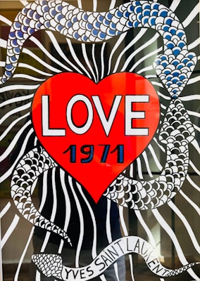 A vibrant illustration features a large red heart with the word 'LOVE' and the year '1971' prominently displayed in bold white lettering. The background includes a black and white striped pattern intertwined with a snake-like design made up of blue and white scales. At the bottom, a ribbon with the text 'YVES SAINT LAURENT' is wrapped around the design.
