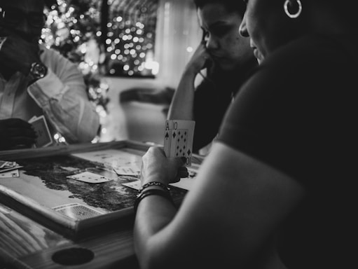 Three people are engaged in a card game, sitting around a table in a cozy, dimly lit setting. One player is holding an Ace and a Ten of diamonds. There are fairy lights and a blurred Christmas tree in the background, adding a festive and warm atmosphere to the scene.