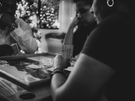 Three people are engaged in a card game, sitting around a table in a cozy, dimly lit setting. One player is holding an Ace and a Ten of diamonds. There are fairy lights and a blurred Christmas tree in the background, adding a festive and warm atmosphere to the scene.