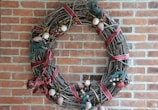 A decorative wreath made of intertwined branches and adorned with red ribbons, artificial fruits, and foliage. It is hung on a brick wall, showcasing a rustic and festive appearance.