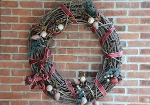 A decorative wreath made of intertwined branches and adorned with red ribbons, artificial fruits, and foliage. It is hung on a brick wall, showcasing a rustic and festive appearance.