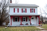 A two-story house with white siding and red accents, including shutters and porch trim. The porch is adorned with various items such as a bucket and a small bicycle. Leafless trees surround the house, indicating a late autumn or winter setting. A small shed and a parked vehicle are visible in the left background.