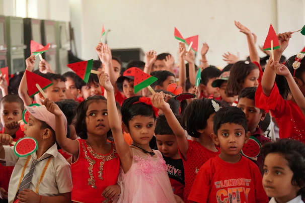 A group of young children in festive attire holding up paper cutouts of watermelons. The children are predominantly wearing red, and many are looking towards the camera. The atmosphere appears joyful and celebratory, likely part of a school event or classroom activity.