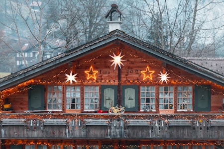 A rustic wooden cabin adorned with festive lights and star decorations. The balcony features traditional elements and is lit up with warm string lights. In the background, leafless trees can be seen, and the cabin's exterior exudes a cozy, holiday charm.