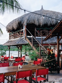 A tropical outdoor restaurant setup featuring wooden tables and chairs with bright red cushions. A thatched roof structure towers above with stairs leading up to an elevated area. A Christmas tree adorned with ornaments and a big bow is visible, adding a festive touch. Palm tree leaves and strands of string lights drape across the setting, with a serene waterfront and lush greenery in the background.