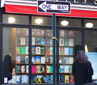 A storefront display features a large assortment of books in a window, with a one-way street sign positioned prominently in the foreground. The books are arranged neatly in rows, showcasing various colorful covers and titles. A person with long hair is standing in front of the window, partially visible from the back.