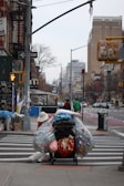 A person is seen pushing a heavily loaded cart across a crosswalk in an urban setting. The cart is filled with large plastic bags, presumably containing recyclable materials like bottles and cans. The background shows city buildings, traffic signals, and signs including a one-way street sign. There is another person dressed in a green jacket walking further down the crosswalk. Various urban elements like a bus stop and a trash bin are also visible.