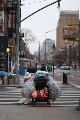 A person is seen pushing a heavily loaded cart across a crosswalk in an urban setting. The cart is filled with large plastic bags, presumably containing recyclable materials like bottles and cans. The background shows city buildings, traffic signals, and signs including a one-way street sign. There is another person dressed in a green jacket walking further down the crosswalk. Various urban elements like a bus stop and a trash bin are also visible.