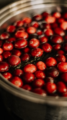 A close-up view of a metal pot filled with fresh cranberries submerged in water. The berries have a glossy texture and a deep red color, contrasting with the silver of the pot.