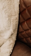 A close-up view of a textured fabric and a quilted leather surface. The fabric appears soft and fluffy, suggesting a warm and cozy material, while the leather surface has a rich brown tone with visible stitching in a diamond pattern.