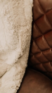 A close-up view of a textured fabric and a quilted leather surface. The fabric appears soft and fluffy, suggesting a warm and cozy material, while the leather surface has a rich brown tone with visible stitching in a diamond pattern.