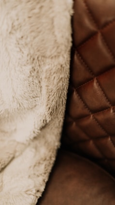 A close-up view of a textured fabric and a quilted leather surface. The fabric appears soft and fluffy, suggesting a warm and cozy material, while the leather surface has a rich brown tone with visible stitching in a diamond pattern.