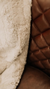A close-up view of a textured fabric and a quilted leather surface. The fabric appears soft and fluffy, suggesting a warm and cozy material, while the leather surface has a rich brown tone with visible stitching in a diamond pattern.
