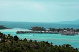 A serene and picturesque view of overwater bungalows surrounded by turquoise blue waters and lush greenery. The ocean stretches into the horizon, blending with the clear sky. Palm trees and dense foliage add a tropical vibe.