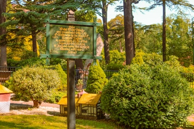 A green historical sign stands in a lush garden surrounded by various shrubs and trees. The sign contains gold text detailing the history of a miniature village presented to a group in England. The background features a fence and more greenery, creating a serene and picturesque setting.